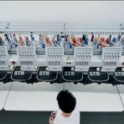 Wielogłowicowa hafciarka przemysłowa w trakcie wykonywania haftu 'STR' na czarnych elementach odzieży, widok z góry na stanowisko pracy.