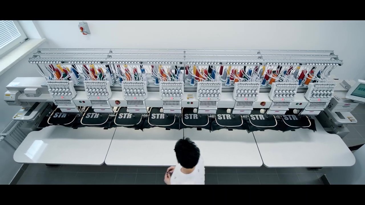 Wielogłowicowa maszyna hafciarska w trakcie pracy nad serią czarnych toreb sportowych z białym napisem 'STR', widok z góry na pracownika obsługującego urządzenie.