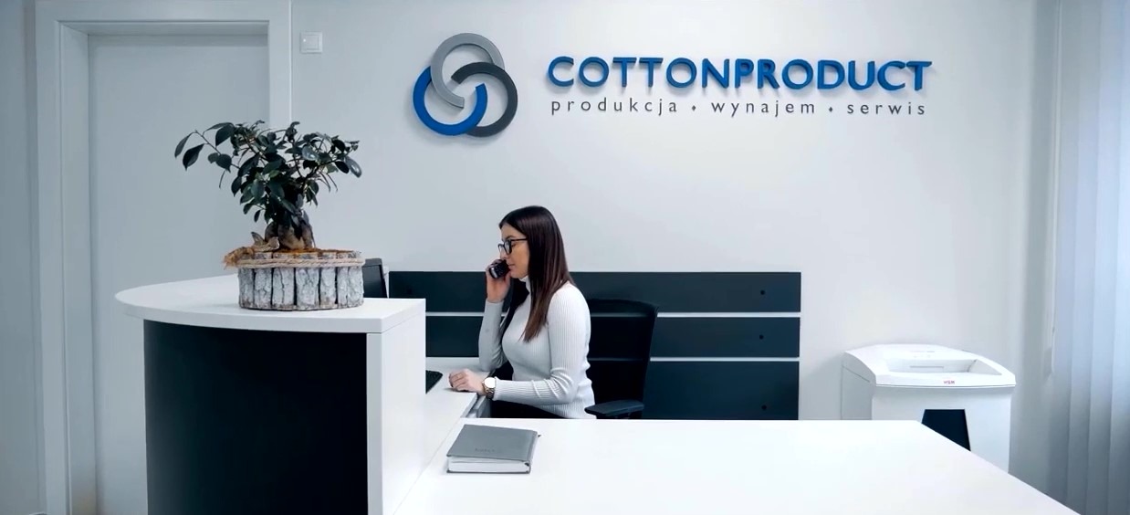 Recepcja firmy Cottonproduct z logo, kobieta w okularach rozmawia przez telefon, na biurku roślina bonsai i notes.