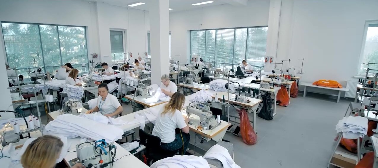 Jasne, przestronne wnętrze szwalni z rzędami stanowisk pracy, gdzie krawcowe szyją na maszynach, stosy białych materiałów na stołach, duże okna z żaluzjami w tle.