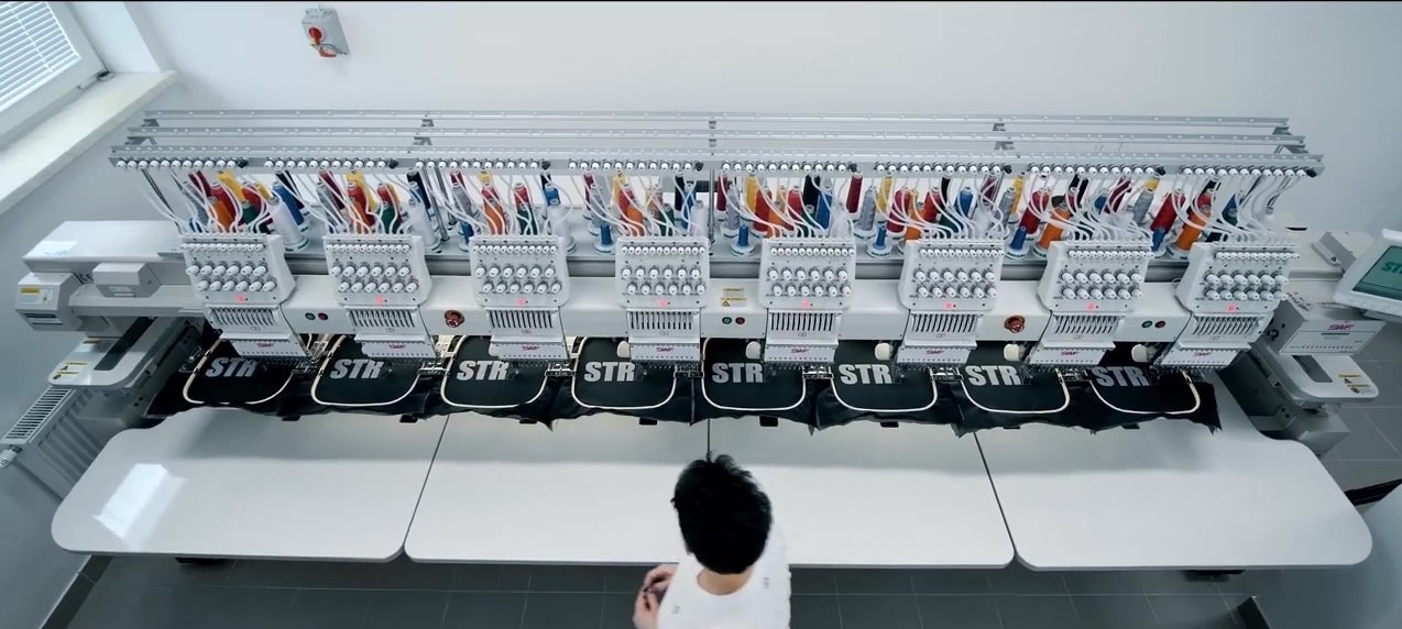 Wielogłowicowa hafciarka przemysłowa w trakcie wykonywania haftu 'STR' na czarnych elementach odzieży, widok z góry na stanowisko pracy.