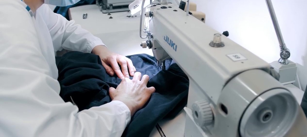 Pracownik w białym fartuchu zszywa ciemny materiał na profesjonalnej maszynie do szycia JUKI w jasnym pomieszczeniu.