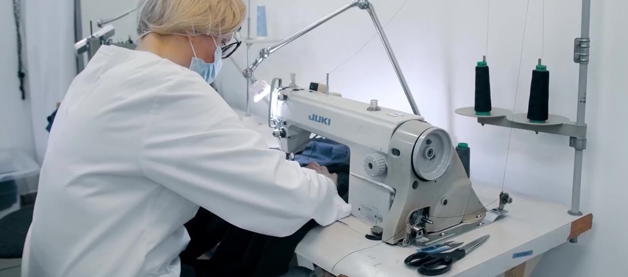 Pracownica w maseczce i czepku ochronnym szyje na maszynie JUKI, obok stojak z czarnymi nićmi, na stole nożyczki i obcinaczki.