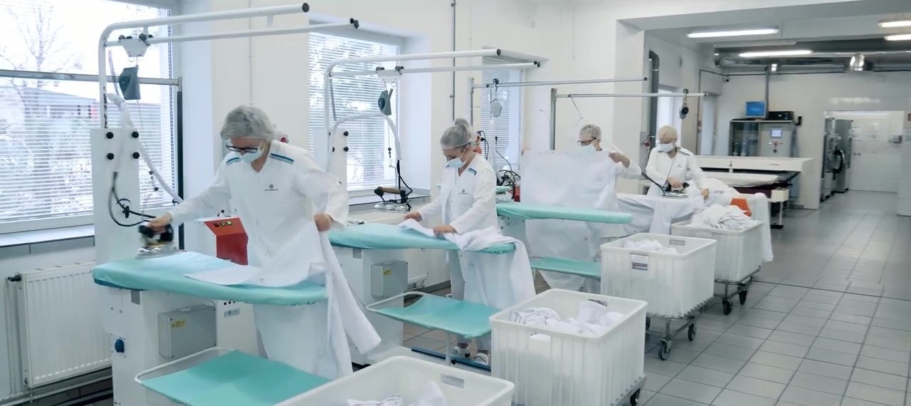 Pracownicy w białych uniformach prasują białą odzież na profesjonalnych stołach prasowalniczych w jasnej, przestronnej pralni przemysłowej.