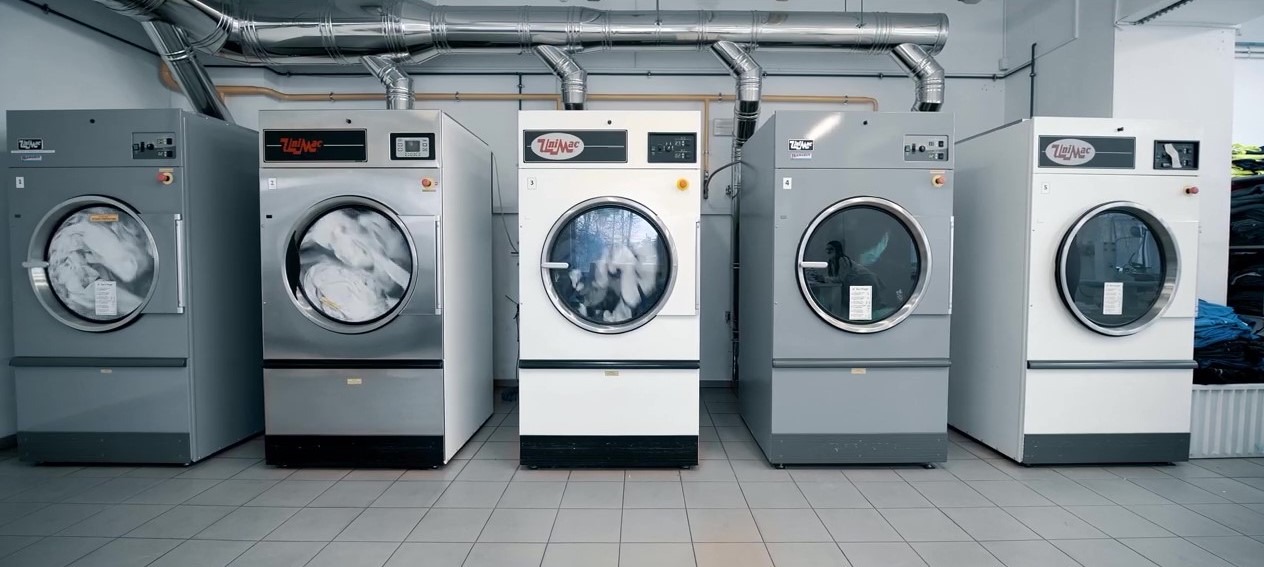Rząd przemysłowych pralek Unimac w szaro-białej kolorystyce, niektóre z wirującym praniem wewnątrz bębnów, rury wentylacyjne nad urządzeniami.