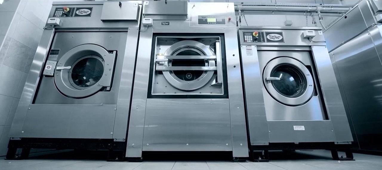 Trzy profesjonalne pralki przemysłowe ze stali nierdzewnej, ustawione obok siebie w pralni, z widocznymi panelami sterowania i drzwiami bębnowymi.