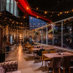 Elegancka restauracja z przeszklonymi ścianami, oświetlona girlandami żarówek, z drewnianymi stolikami i różnokolorowymi krzesłami oraz pluszową sofą w rogu.