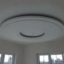 Okrągła, biała dekoracja sufitowa z wgłębieniem na lampę, widoczna w rogu pokoju z oknami, ściany przygotowane do malowania.