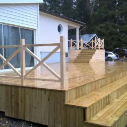 Świeżo wybudowany, drewniany taras z balustradą i schodami, przylegający do jasnego domu z dużymi oknami przesuwnymi, widok na las w tle, mokra powierzchnia po deszczu.