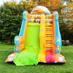 Kolorowa, nadmuchiwana zjeżdżalnia w kształcie zamku na trawniku, z widocznymi schodkami i zjeżdżalnią, otoczona zielonymi drzewami.