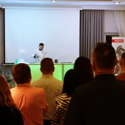 Barman w białym uniformie przygotowuje drinki podczas pokazu dla grupy osób, widoczny podświetlony bar i ekran projekcyjny w tle, oraz baner reklamowy ERKO.