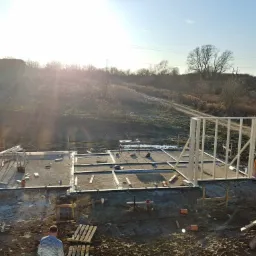 Wczesny etap budowy domu szkieletowego: fundament z drewnianą konstrukcją szkieletową, widok z góry na plac budowy z pracującym mężczyzną i zaparkowanym vanem.