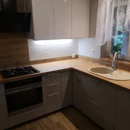 Nowoczesna kuchnia z szarymi szafkami, drewnianym blatem, płytą gazową i piekarnikiem Amica, oświetleniem LED pod szafkami wiszącymi oraz dekoracyjnymi płytkami ściennymi w falisty wzór.