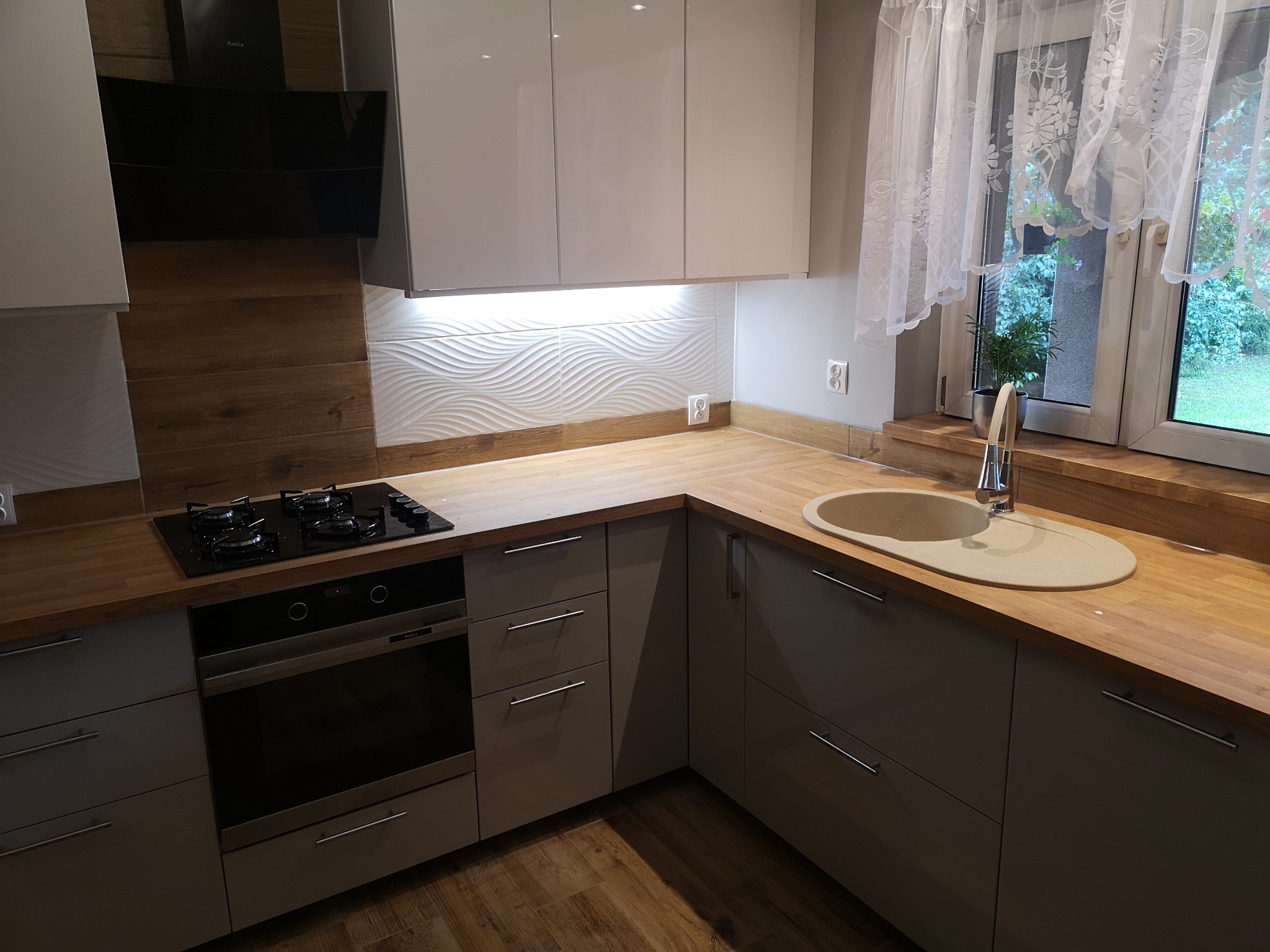 Nowoczesna kuchnia z szarymi szafkami, drewnianym blatem, płytą gazową i piekarnikiem Amica, oświetleniem LED pod szafkami wiszącymi oraz dekoracyjnymi płytkami ściennymi w falisty wzór.