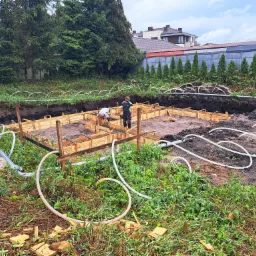 Przygotowanie szalunku pod fundament budynku jednorodzinnego, widoczni dwaj pracownicy w trakcie montażu, teren budowy zarośnięty trawą i chwastami, w tle rząd tuj i budynki mieszkalne.