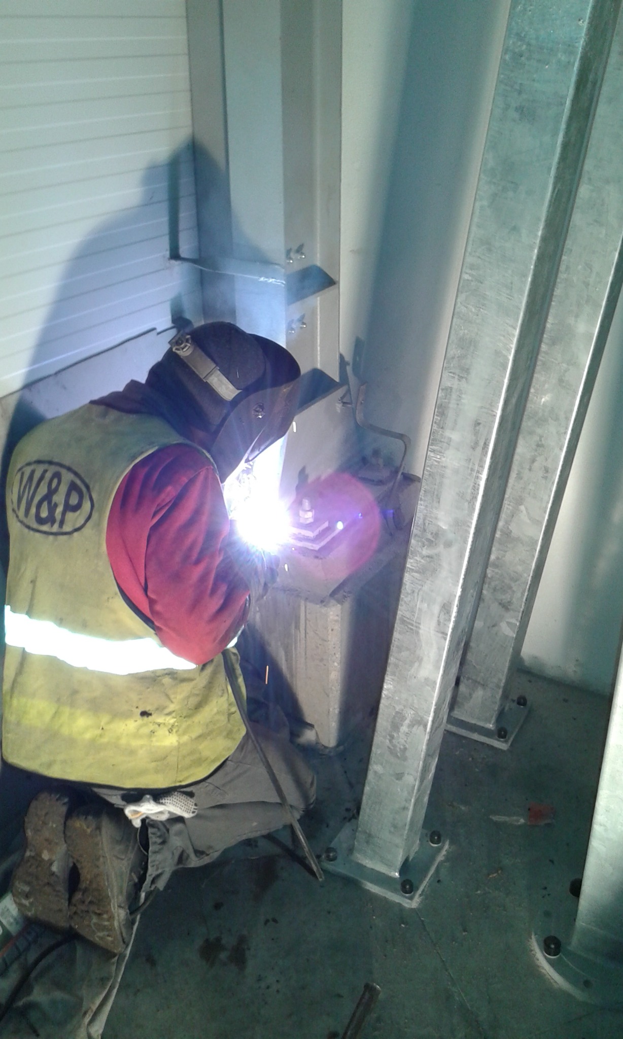 Pracownik w odzieży ochronnej, z maską spawalniczą, wykonuje spawanie metalowej konstrukcji w hali, widoczne iskry i elementy konstrukcyjne.