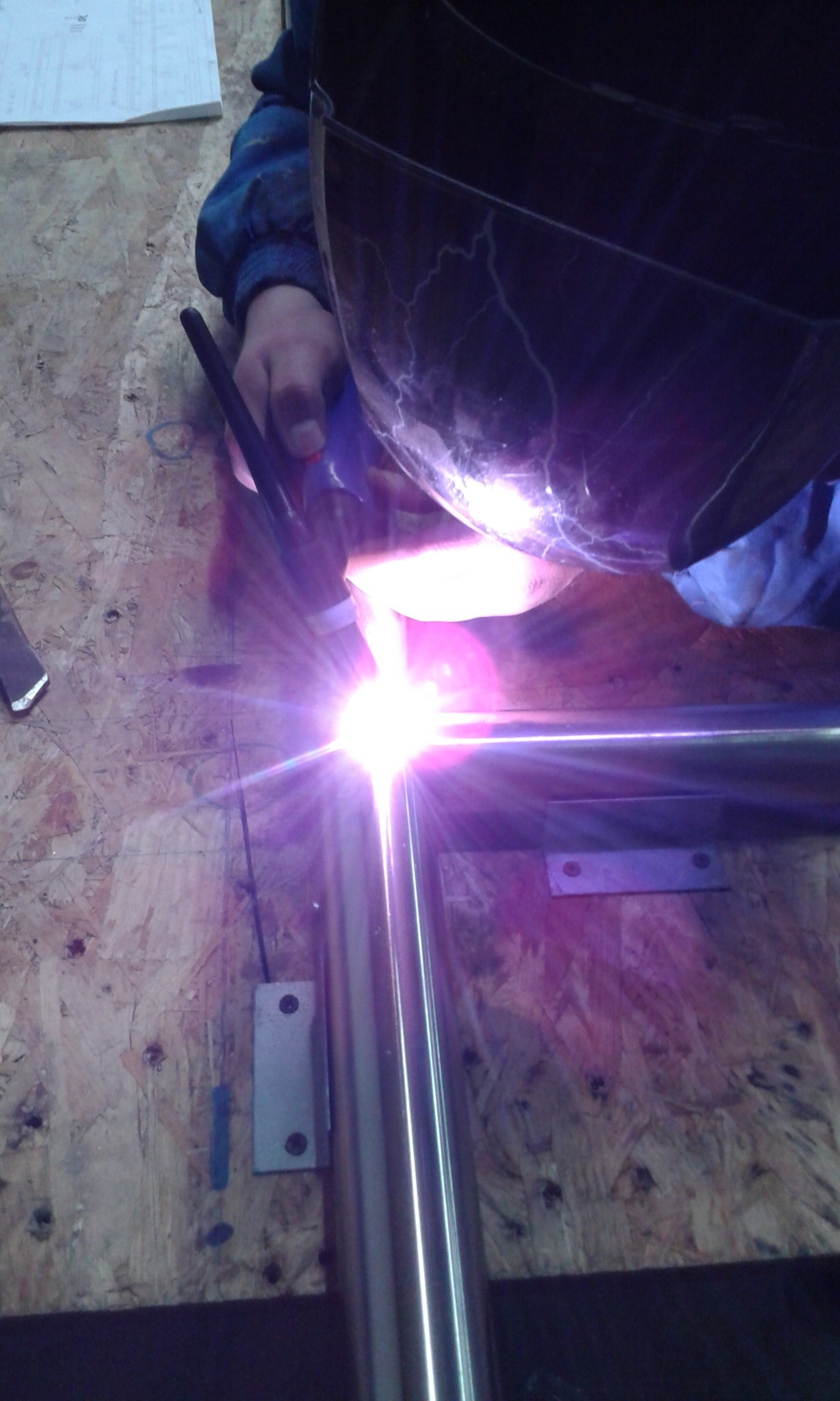 Intensywne światło łuku spawalniczego podczas spawania metalowych rur przez spawacza w masce ochronnej, widoczne iskry i elementy mocujące na drewnianym podłożu.