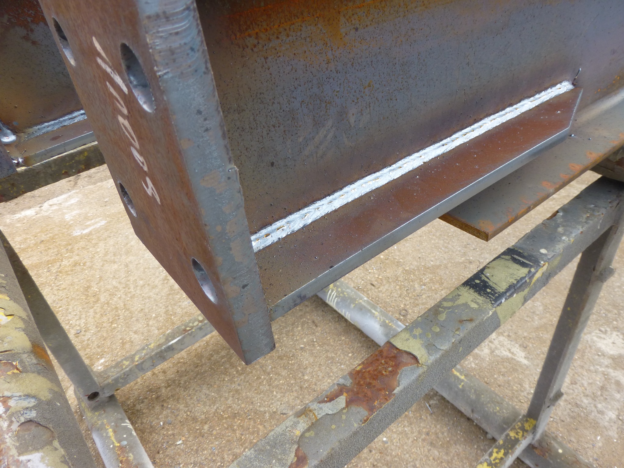 Fragment stalowej belki dwuteowej z widocznym świeżym spoiwem po spawaniu, ustawionej na metalowym stojaku. Na pionowej ściance belki widoczne otwory i białe napisy.