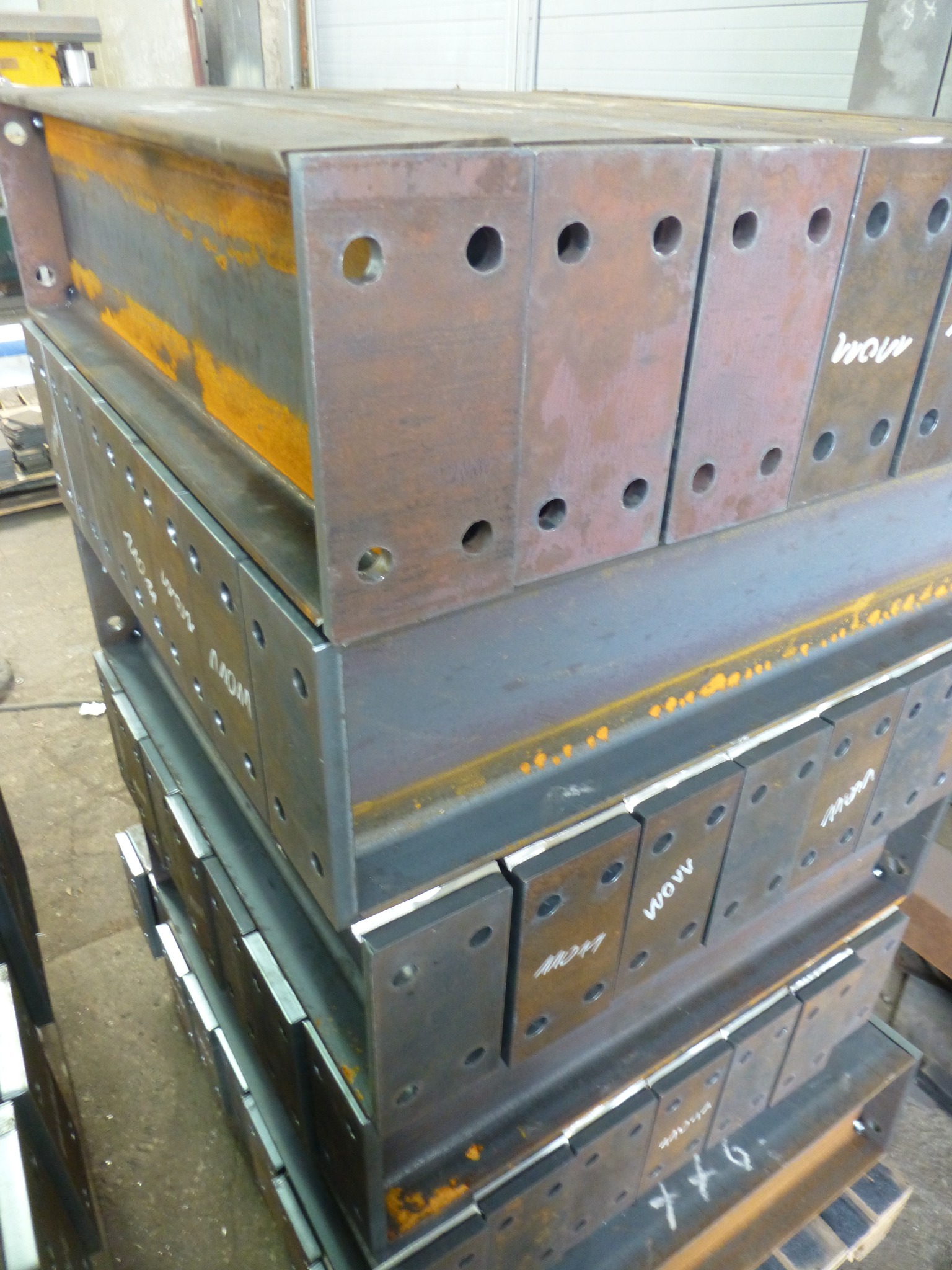 Stos belek stalowych z przyspawanymi płytami wzmacniającymi z otworami, widoczne ślady rdzy i białe napisy na niektórych elementach.