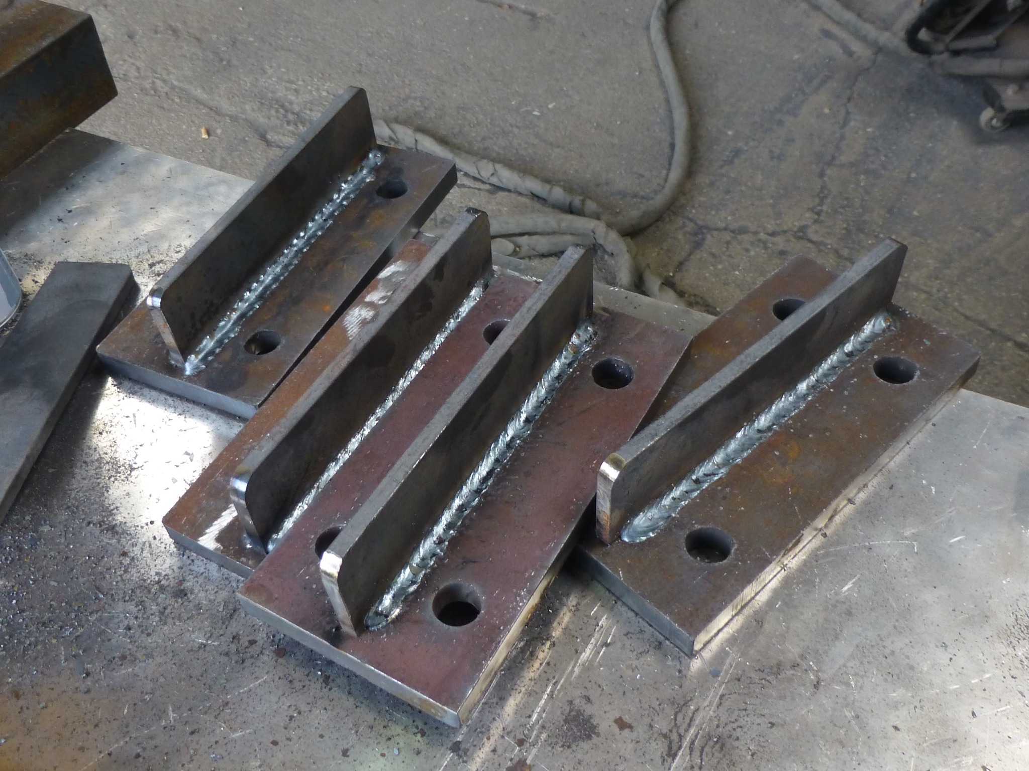 Cztery metalowe elementy konstrukcyjne, zespawane pod kątem prostym, z widocznymi spawami i otworami, leżące na metalowym stole warsztatowym w warsztacie.