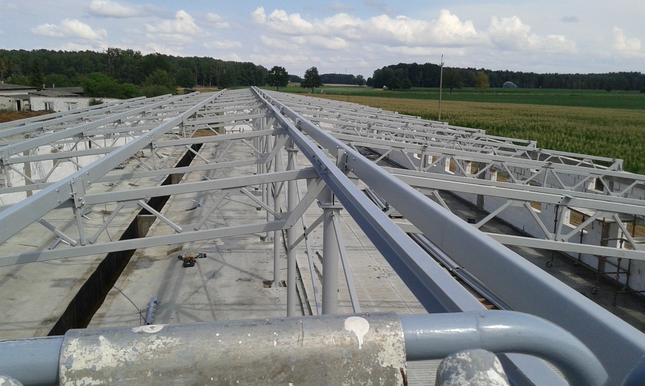 Szkielet dachu hali produkcyjnej z szarej stali, widok z góry na konstrukcję kratownicową i betonowe fundamenty, w tle pola uprawne i las.