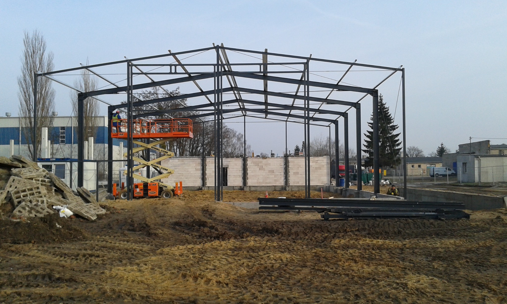 Montaż stalowej konstrukcji hali z użyciem podnośnika koszowego, widoczne elementy konstrukcyjne i fundamenty.