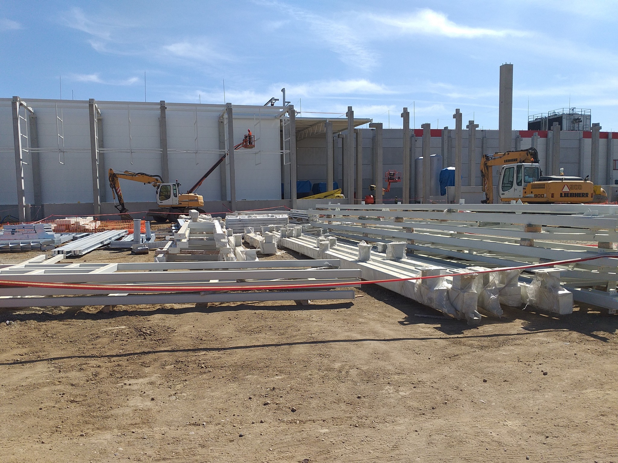 Budowa hali w Obornikach: widok na plac budowy z przygotowanymi elementami konstrukcyjnymi, dwoma koparkami i podnośnikiem koszowym na tle powstającego budynku.