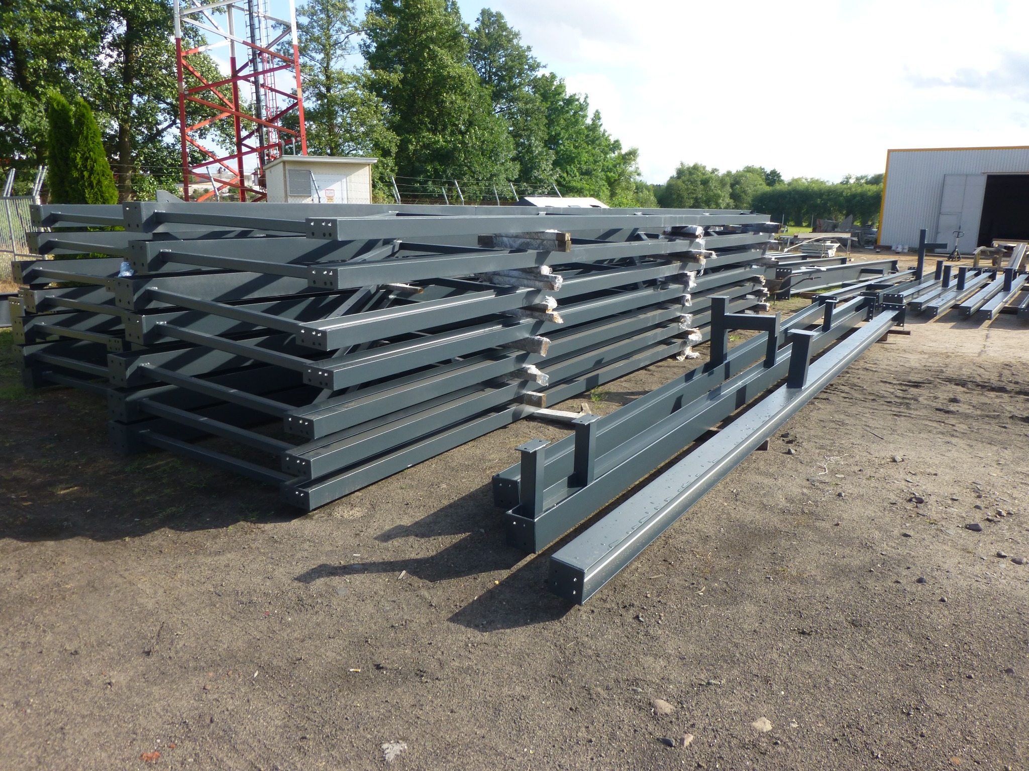 Stos pomalowanych na szaro elementów konstrukcji stalowej z otworami na śruby, przygotowanych do montażu, leżących na ziemi obok hali produkcyjnej