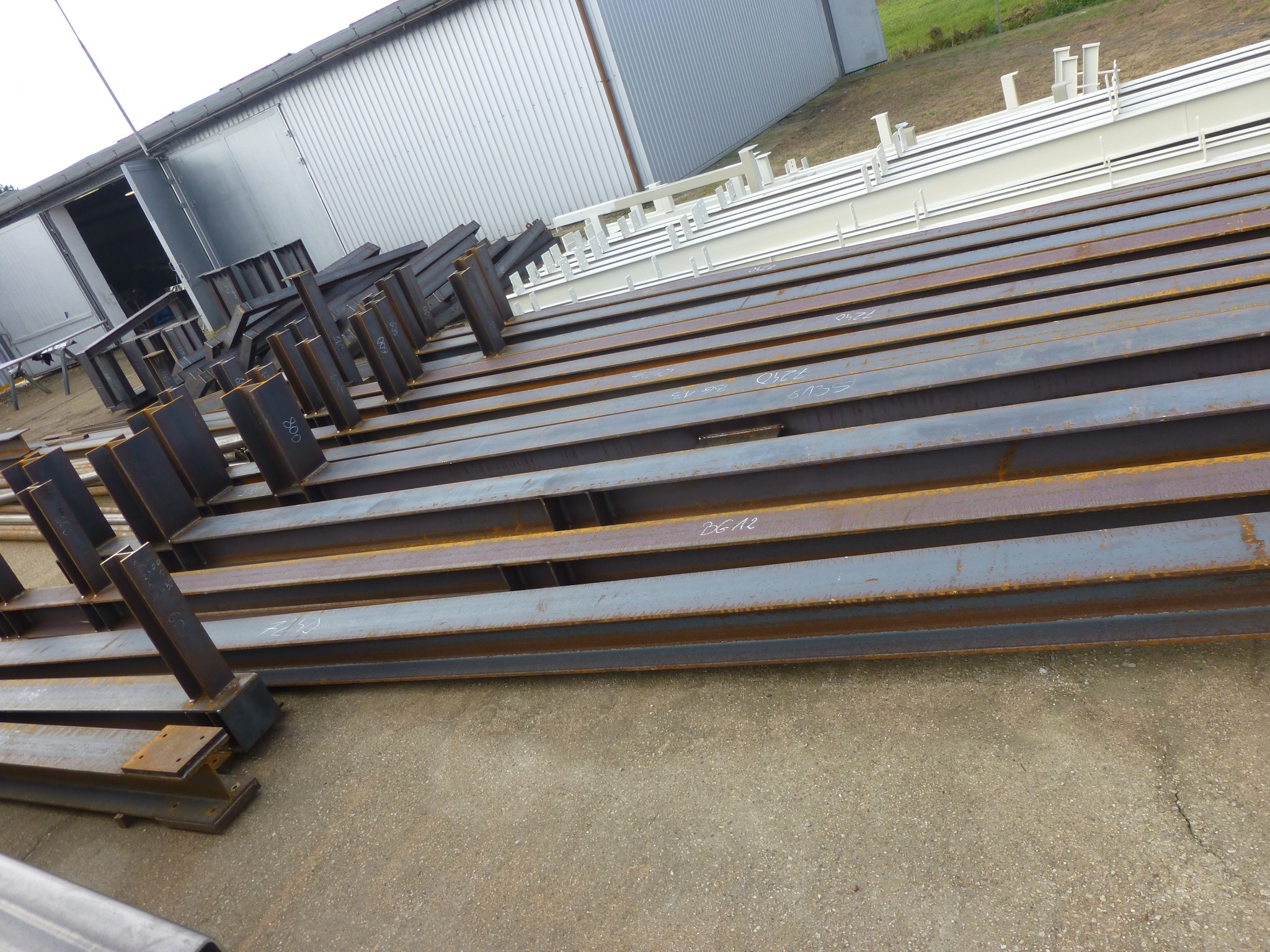 Stalowe belki dwuteowe, niektóre z przyspawanymi elementami, ułożone równolegle na betonowej powierzchni przed halą produkcyjną; niektóre z nich posiadają białe napisy