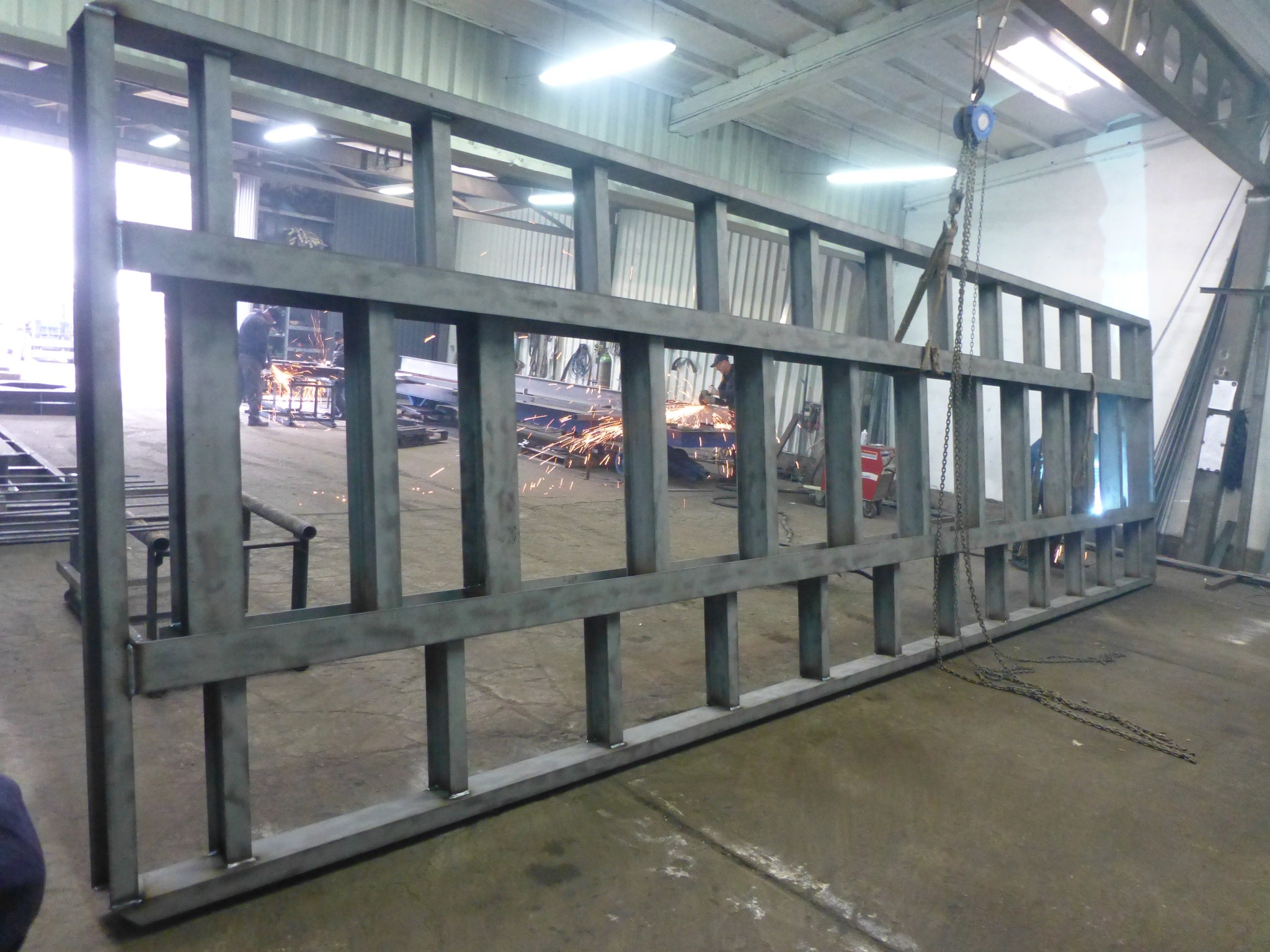 Surowa, spawana rama z profili stalowych, widoczna w hali produkcyjnej, z pracującymi w tle osobami i iskrami szlifierskimi.