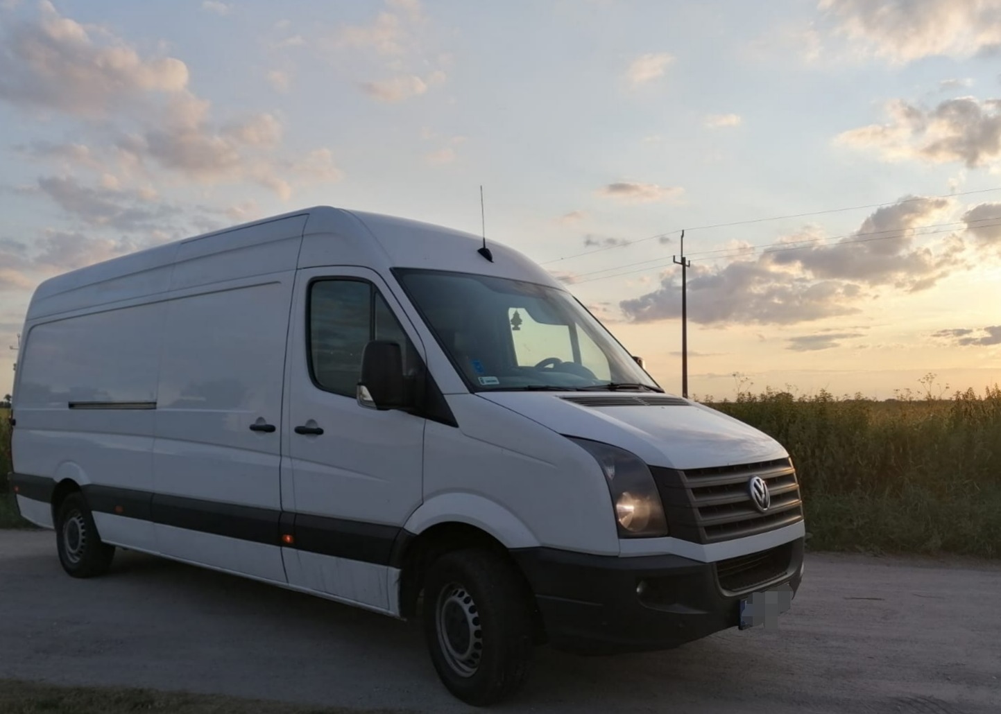 Biały, wysoki bus dostawczy marki Volkswagen Crafter, zaparkowany na polnej drodze na tle zachodzącego słońca i pochmurnego nieba.
