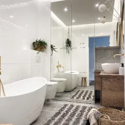 Nowoczesna łazienka z wolnostojącą wanną, umywalką nablatową na drewnianej szafce i dużym lustrem podzielonym na segmenty, podkreślona białymi płytkami i złotymi akcentami armatury.