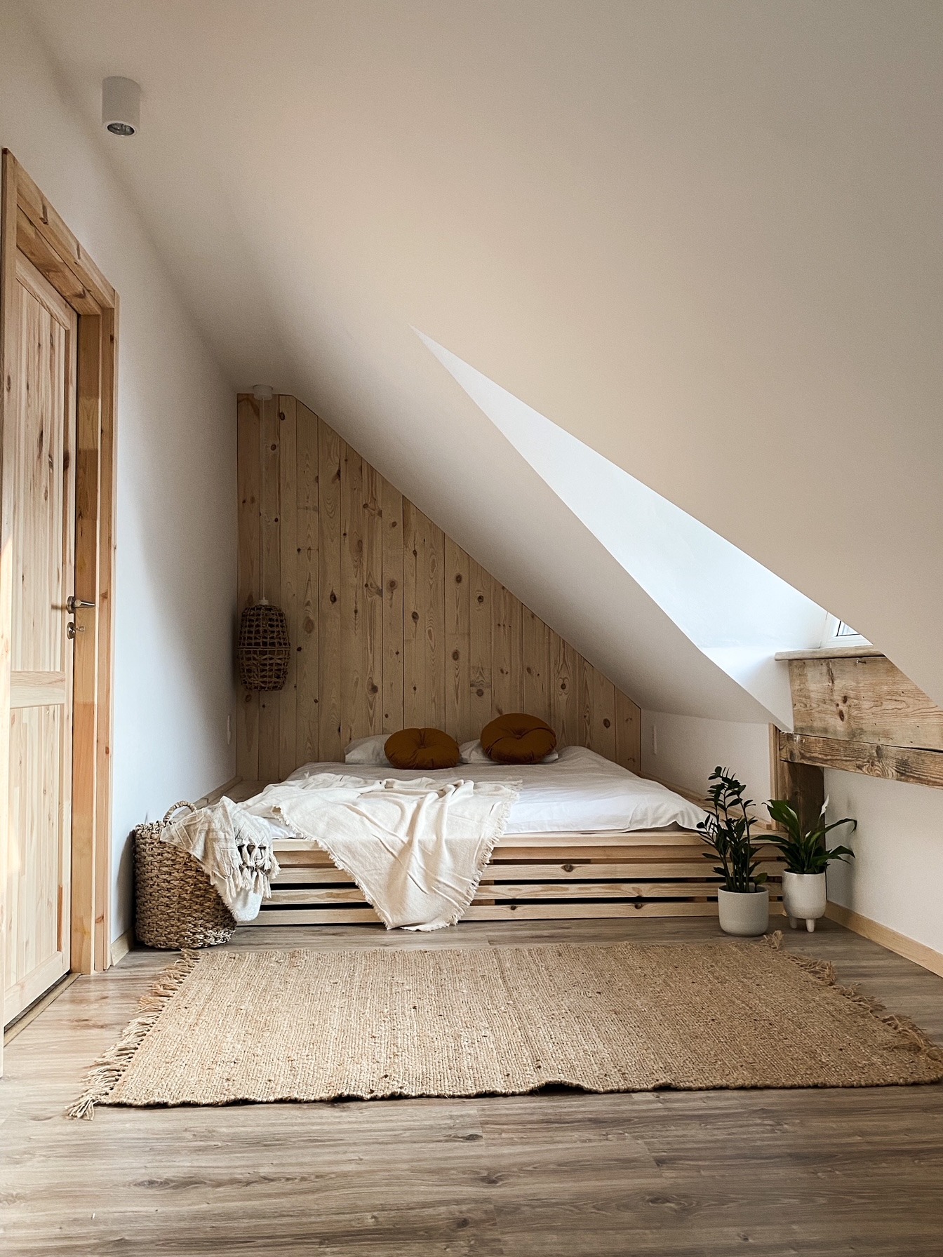 Minimalistyczna sypialnia na poddaszu z drewnianym wykończeniem ściany za łóżkiem, jutowym dywanem i roślinami doniczkowymi, oświetlona przez okno dachowe.