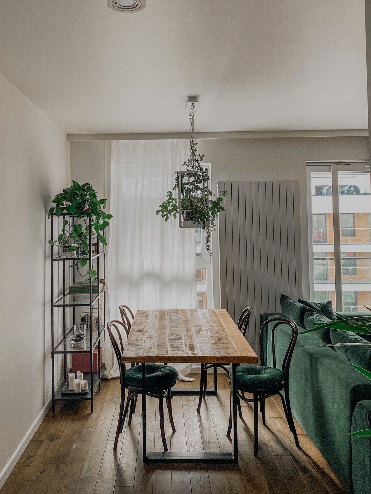 Jasne wnętrze jadalni z drewnianym stołem, krzesłami z zielonymi siedziskami, wiszącą rośliną w geometrycznej donicy i regałem z roślinami.