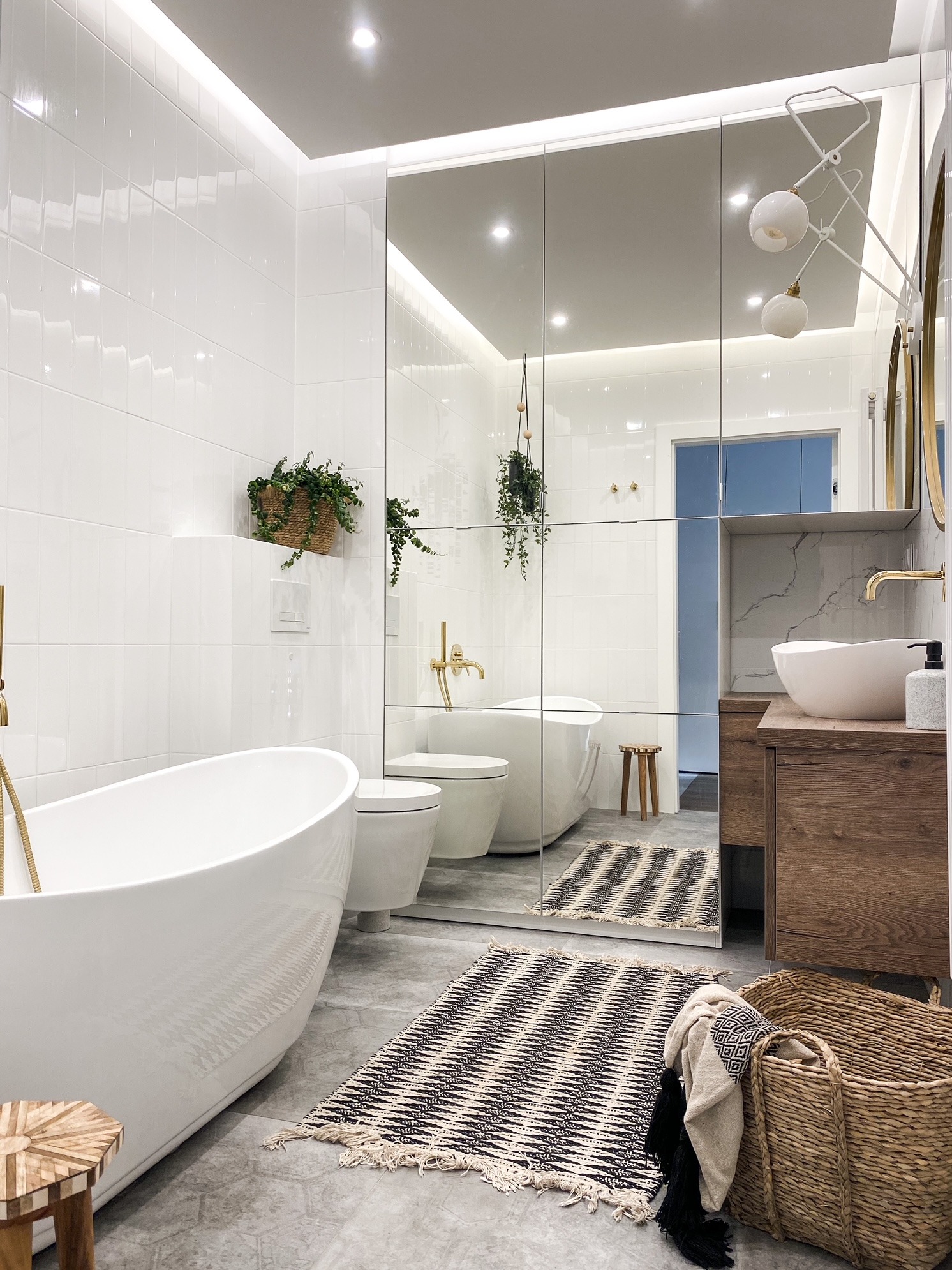 Nowoczesna łazienka z wolnostojącą wanną, umywalką nablatową na drewnianej szafce i dużym lustrem podzielonym na segmenty, podkreślona białymi płytkami i złotymi akcentami armatury.