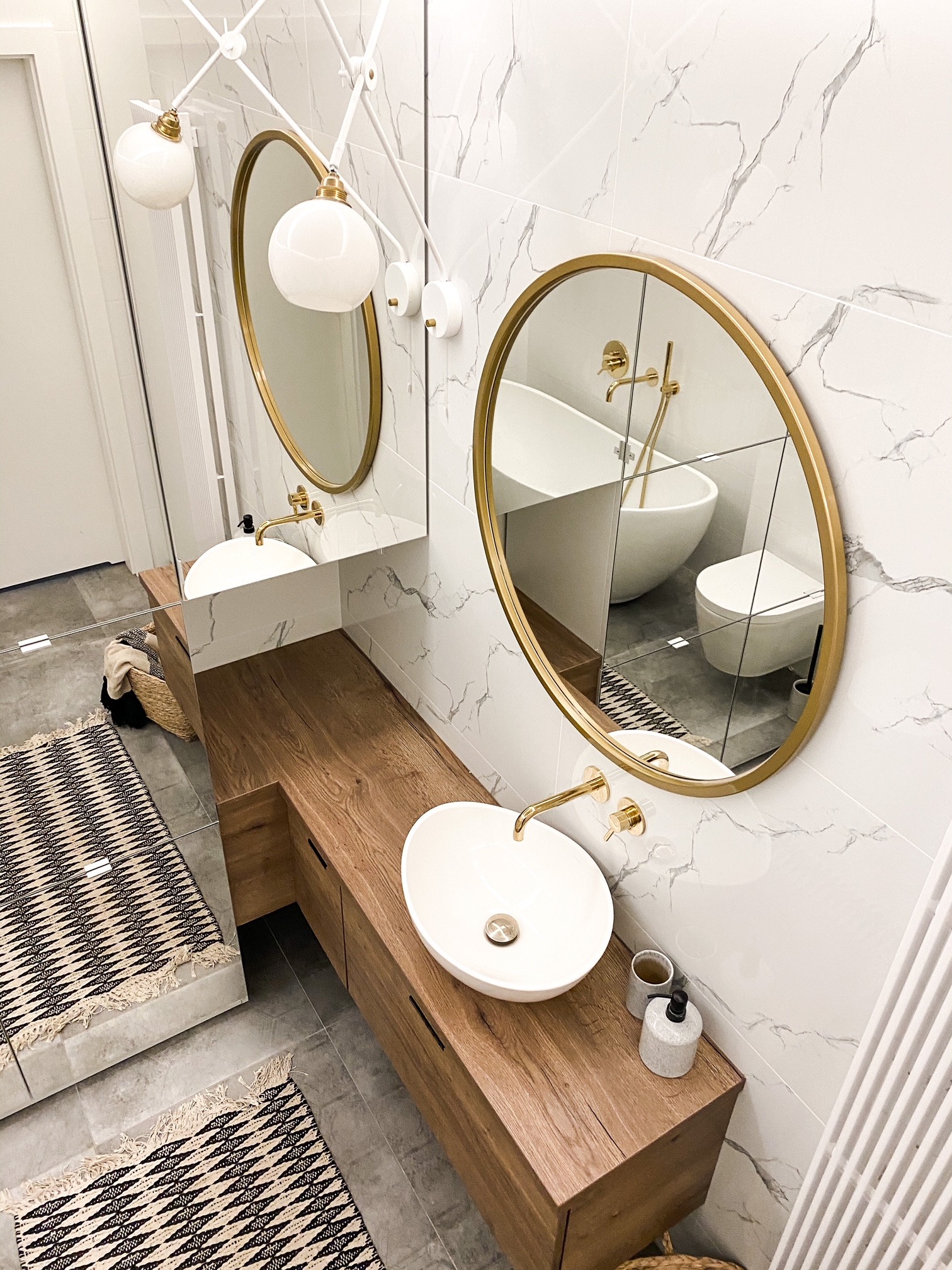 Nowoczesna łazienka z umywalką nablatową w kształcie misy, drewnianą szafką pod umywalkę, okrągłymi lustrami w złotych ramach i białymi płytkami imitującymi marmur na ścianach. W lustrze odbija się...