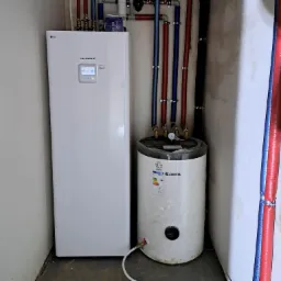 Biała pompa ciepła LG Therma V z podłączonym zasobnikiem ciepłej wody Kospel SVK-100, widoczne rury instalacyjne w kolorach niebieskim i czerwonym, zamontowane w pomieszczeniu technicznym.