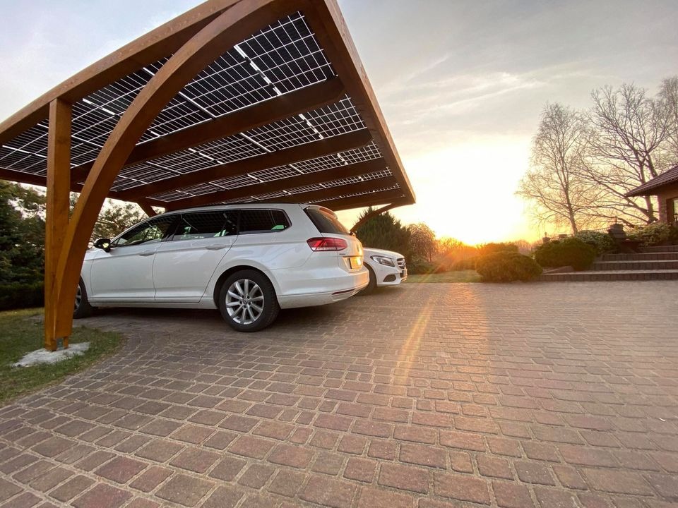 Drewniana wiata solarna z panelami fotowoltaicznymi nad dwoma samochodami osobowymi, kostka brukowa na podjeździe w promieniach zachodzącego słońca.