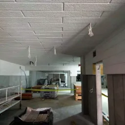 Surowy strop z widoczną fakturą imitującą cegłę, wiszące przewody elektryczne, plac budowy w tle z materiałami i rusztowaniami, żółta taśma ostrzegawcza.