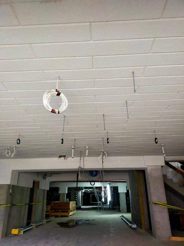 Betonowy sufit z widocznymi hakami i przewodami elektrycznymi w trakcie instalacji, w tle widoczne elementy konstrukcyjne i przejście do kolejnego pomieszczenia.