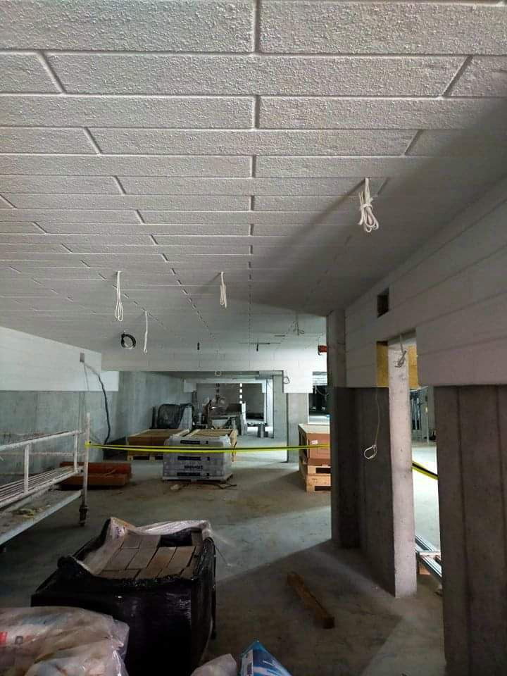 Surowy strop z widoczną fakturą imitującą cegłę, wiszące przewody elektryczne, plac budowy w tle z materiałami i rusztowaniami, żółta taśma ostrzegawcza.