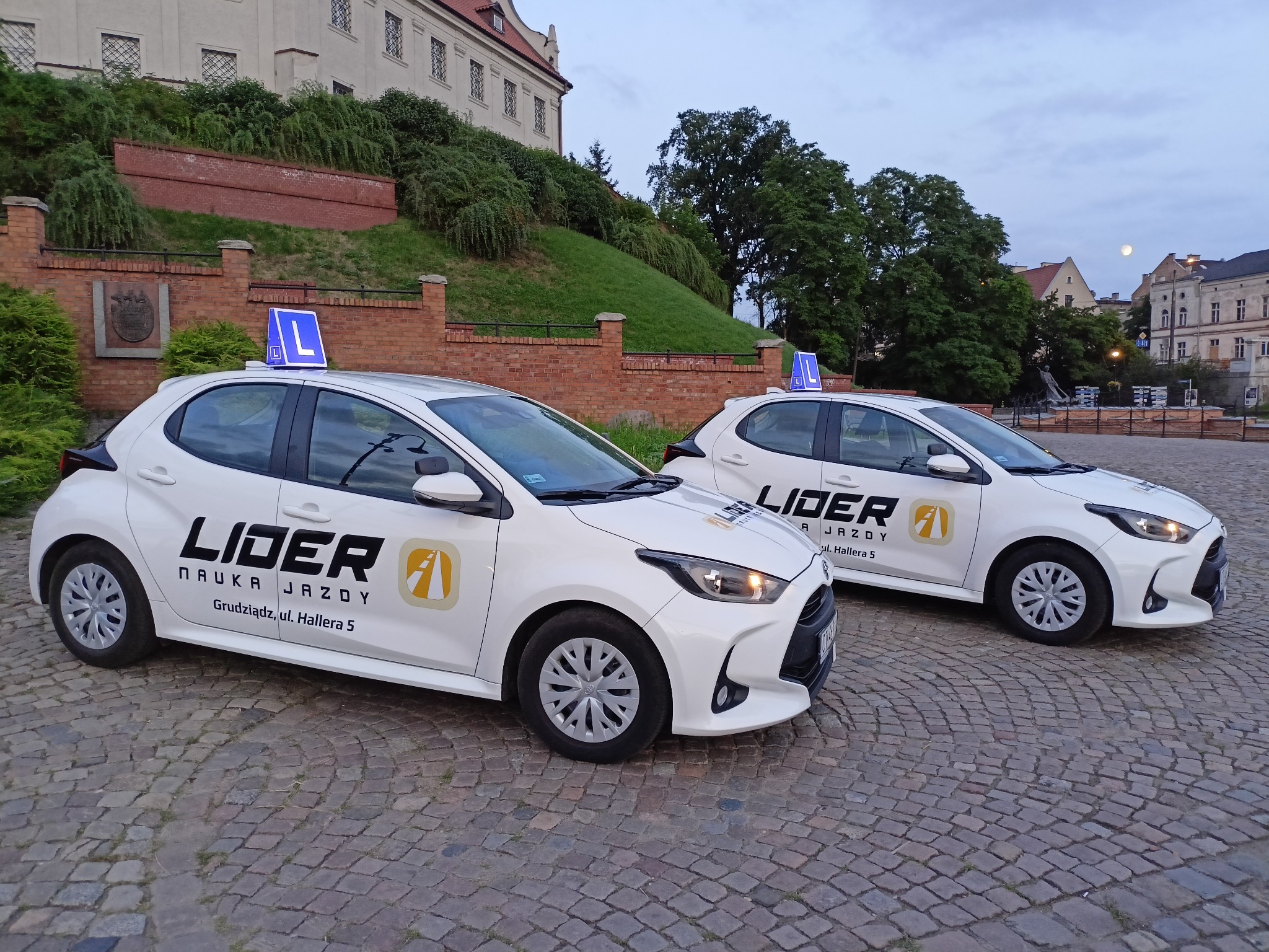 Dwa białe samochody z oznaczeniem 'L' na dachu, zaparkowane na brukowanym placu przed budynkiem z czerwonej cegły i zielonym wzgórzem w tle, widoczny księżyc na niebie.