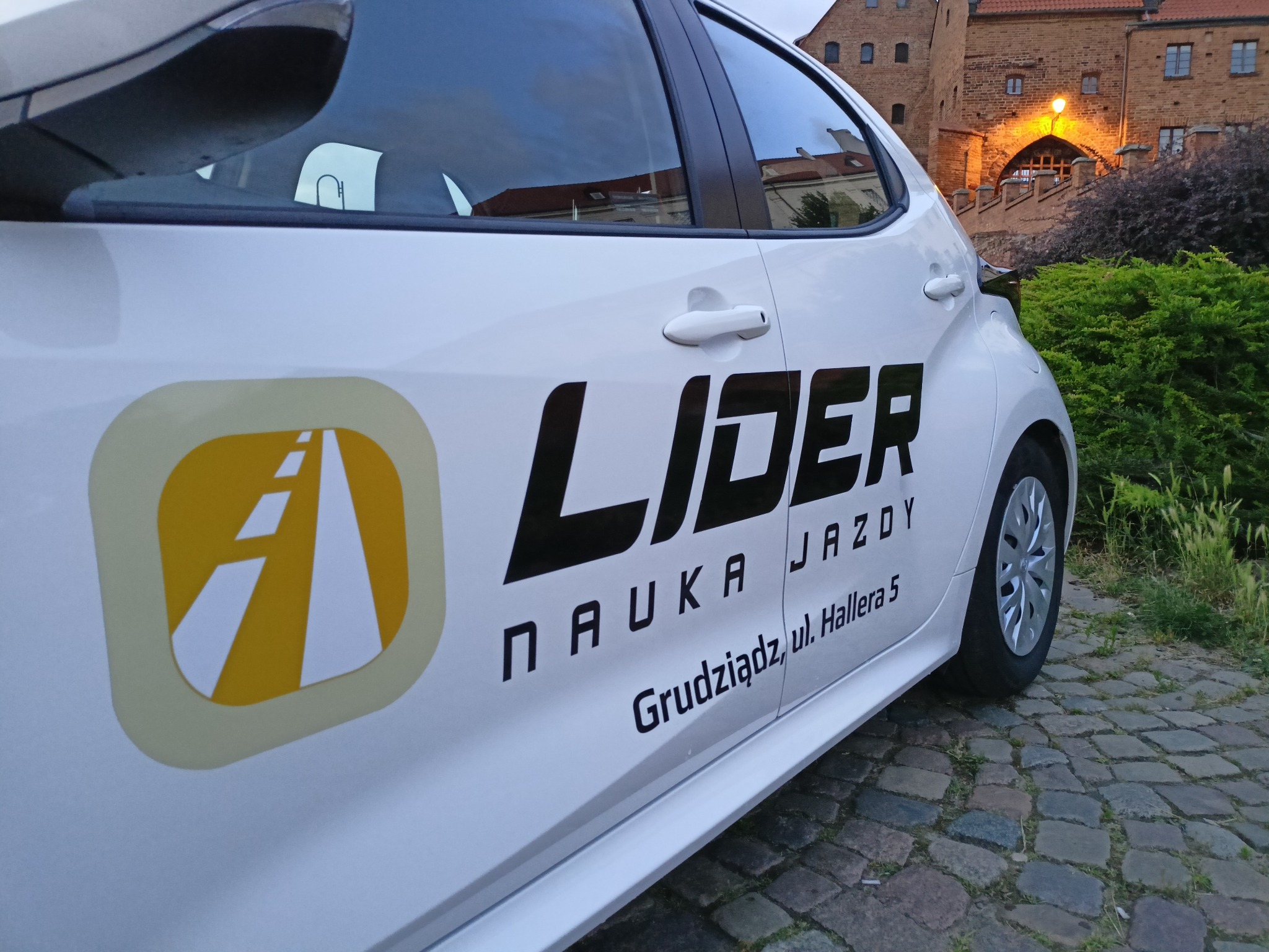 Biały samochód z logotypem szkoły jazdy Lider Nauka Jazdy Grudziądz na tle brukowanej ulicy i ceglanego budynku z bramą.