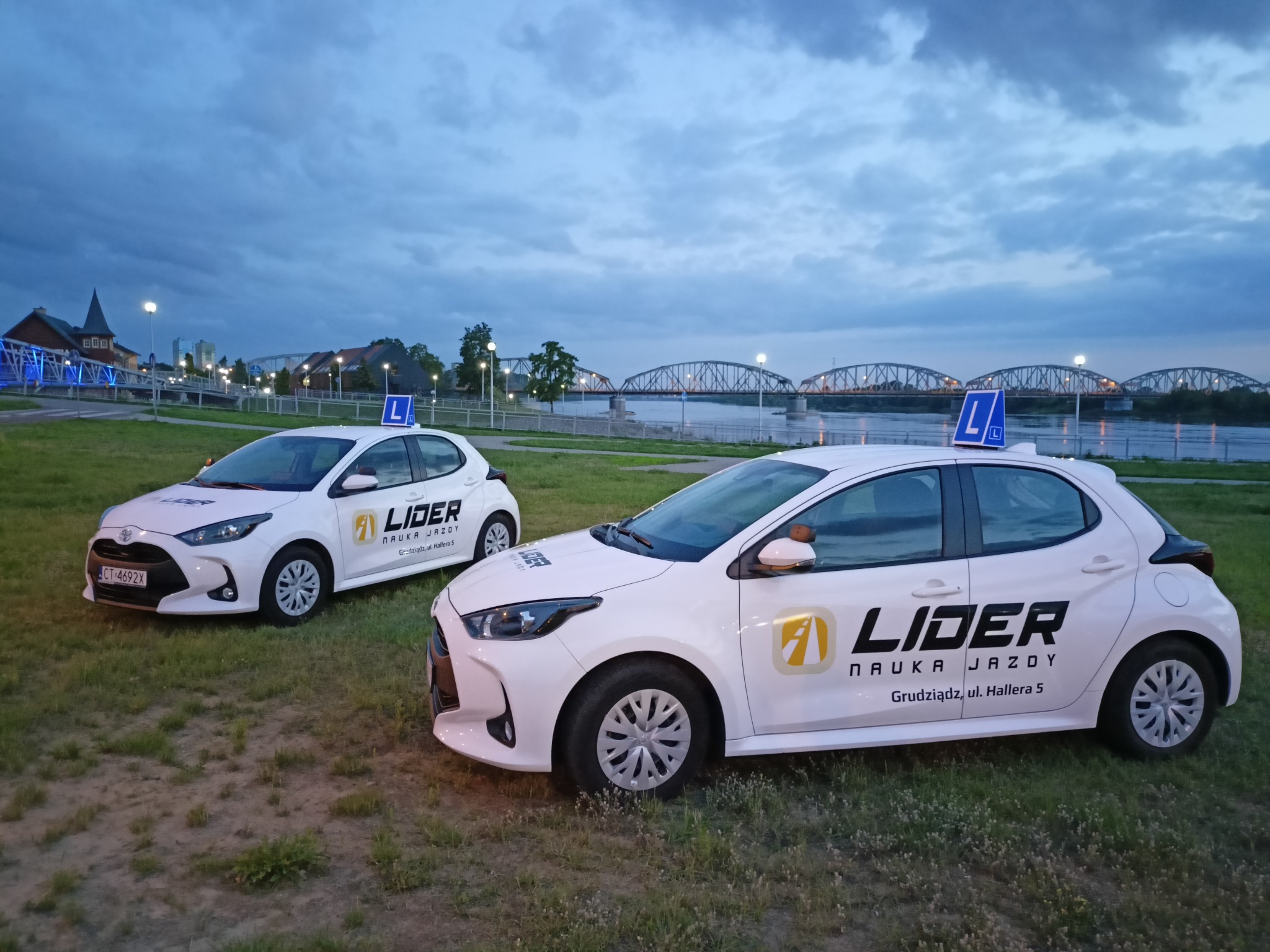 Dwa białe samochody z oznaczeniami szkoły nauki jazdy Lider na tle mostu i rzeki w Grudziądzu, widoczne tablice 'L' na dachu.