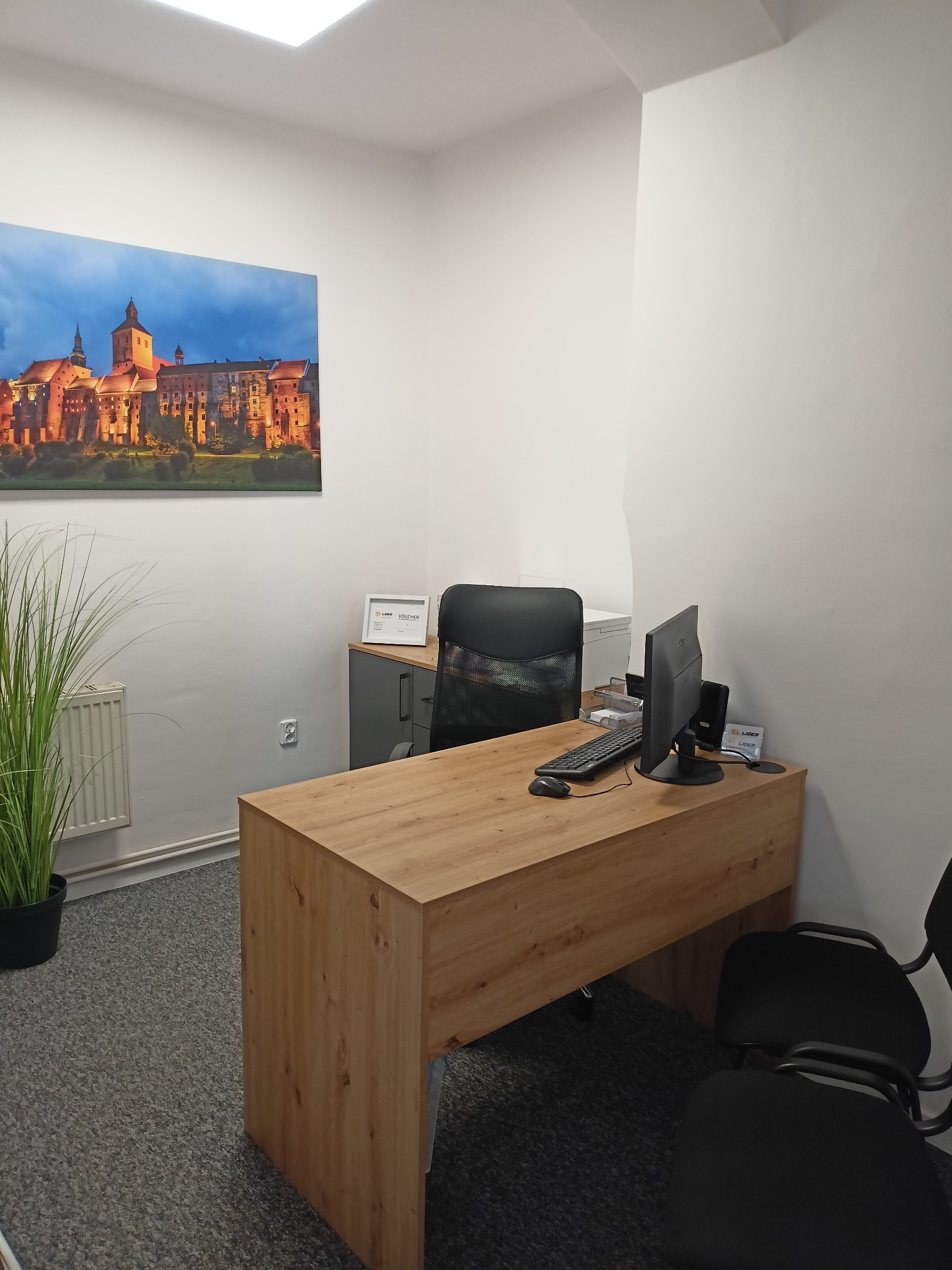 Biuro z drewnianym biurkiem, komputerem, krzesłem biurowym i obrazem zamku na ścianie. Na biurku widoczne dokumenty i voucher.