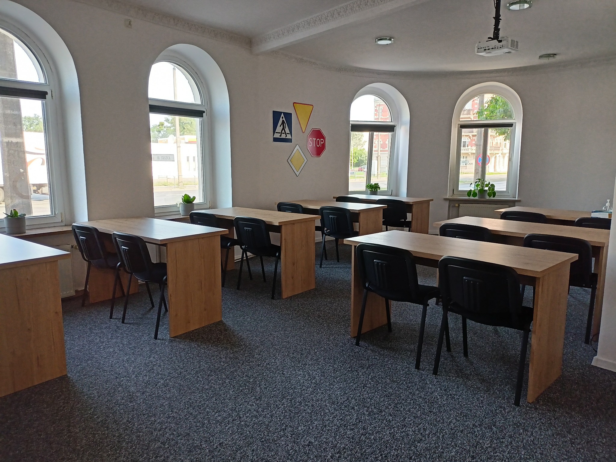 Sala wykładowa z drewnianymi biurkami i czarnymi krzesłami, na ścianie znaki drogowe, rzutnik pod sufitem, okna z roślinami.