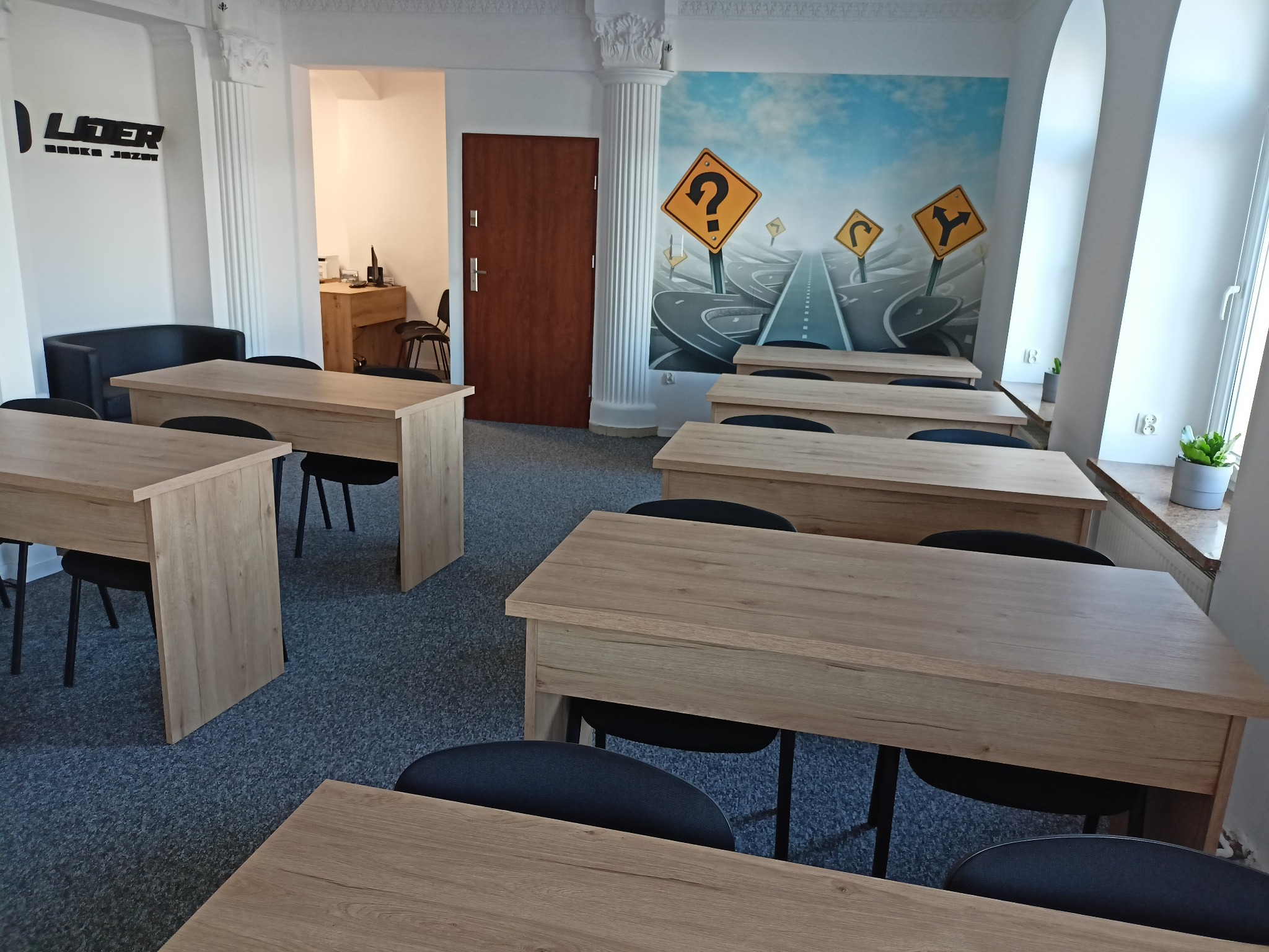 Sala wykładowa z drewnianymi ławkami i czarnymi krzesłami, na ścianie grafika z drogowskazami i znakami zapytania, logo firmy na ścianie.