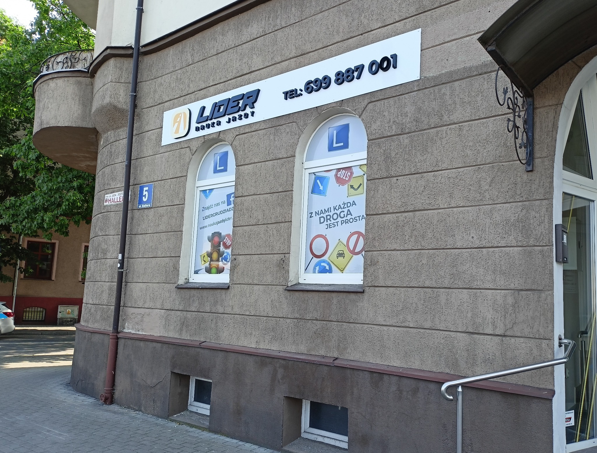 Fasada budynku z szyldem i reklamami w oknach, informującymi o ofercie kursów nauki jazdy, adres na tabliczce.