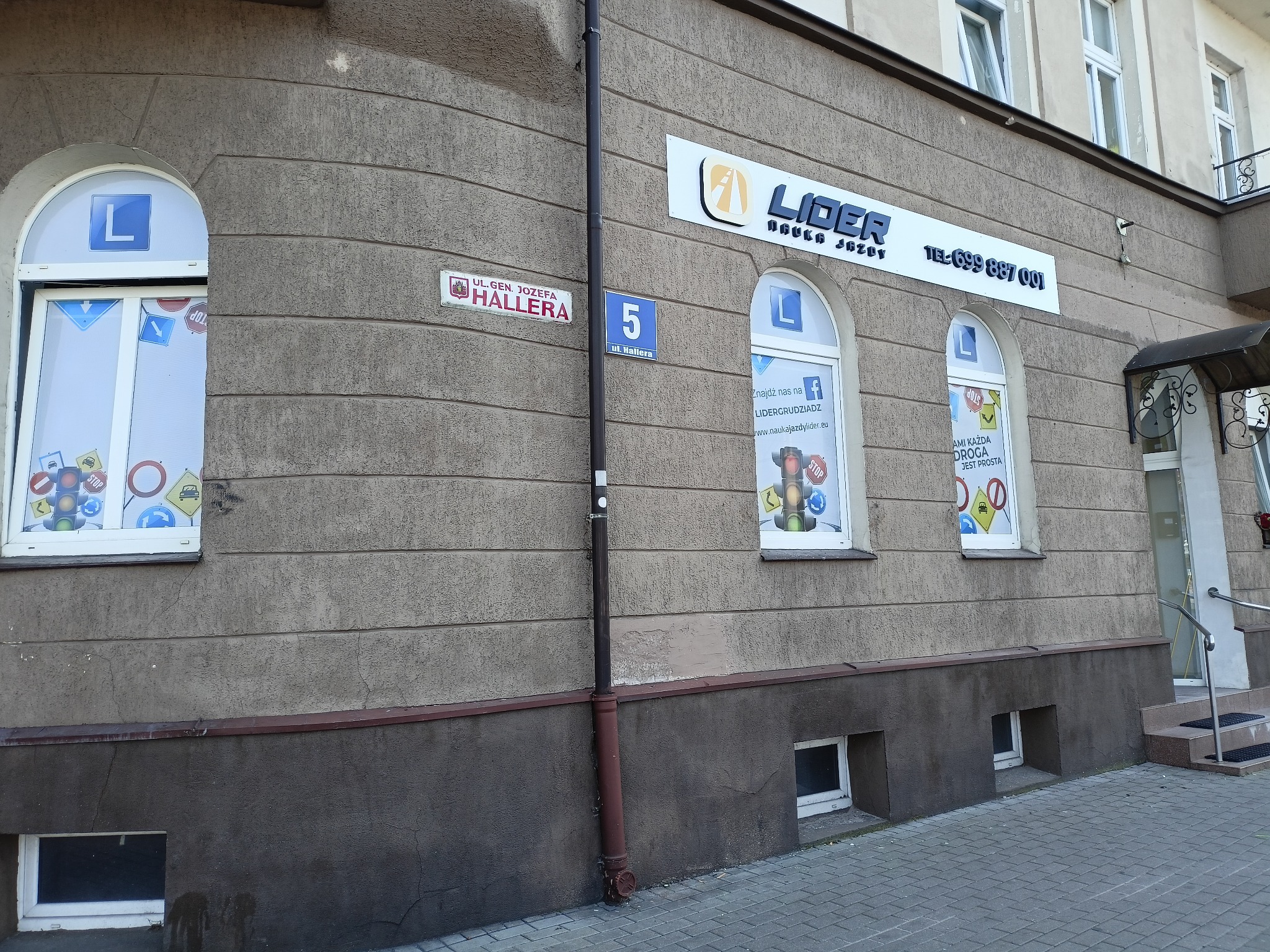 Fasada budynku z szyldem i oknami z naklejkami znaków drogowych, tabliczka z adresem Ul. Gen. Józefa Hallera 5.