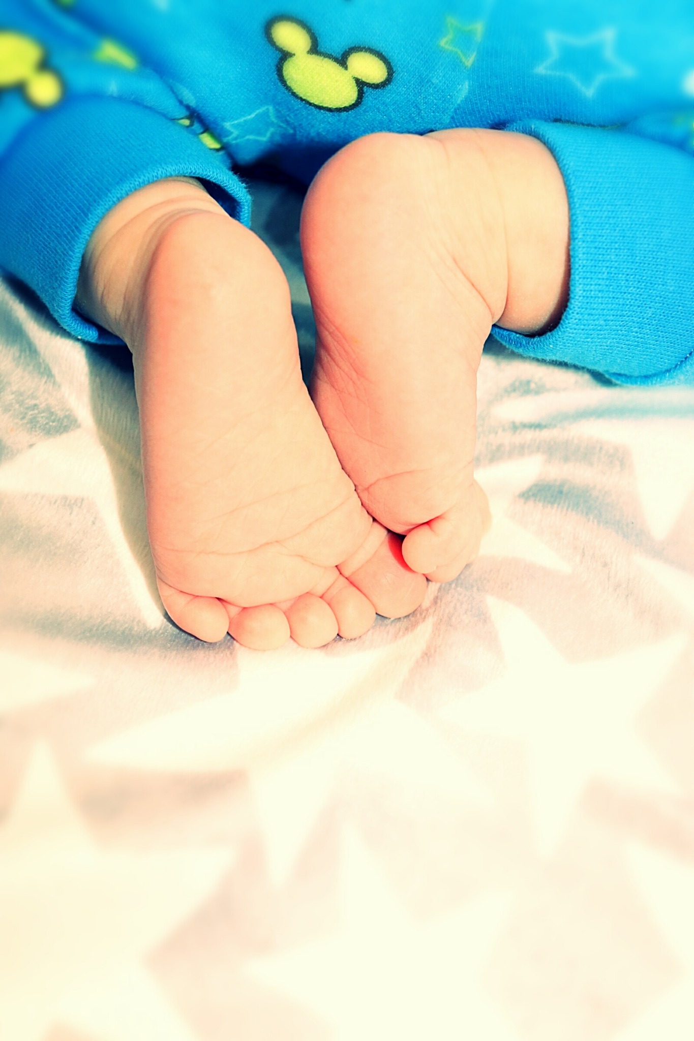 Dwie małe stópki noworodka w niebieskim ubranku z motywem Myszki Miki, leżące na białym kocyku w szare gwiazdki.